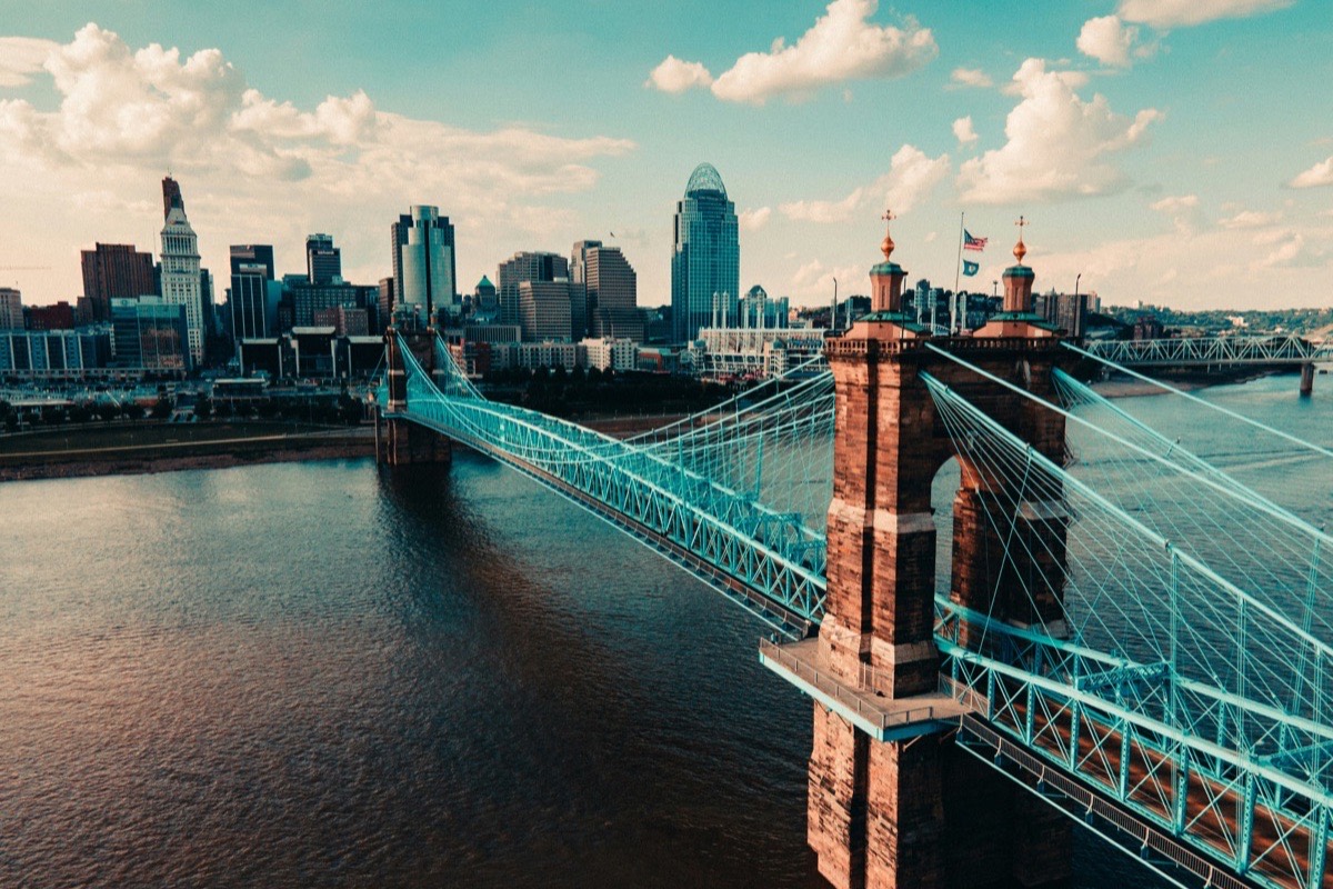 The blue Roebling Suspension Bridge crossing the Ohio River into downtown Cincinnati, with Great American Tower and the Carew Tower rising behind it under a partly cloudy sky