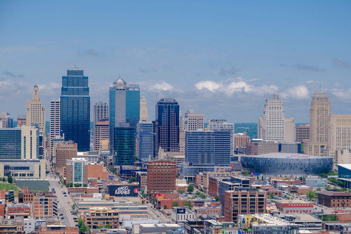 The downtown Kansas City, Missouri skyline on a clear day, with blue glass towers rising above red brick buildings and the round T-Mobile Center arena on the right