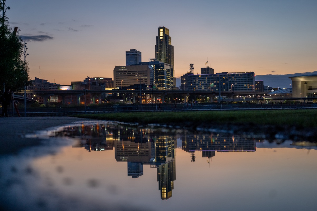 The downtown Omaha, Nebraska skyline at dusk, with the First National Bank Tower lit up and the buildings reflected in a puddle in the foreground