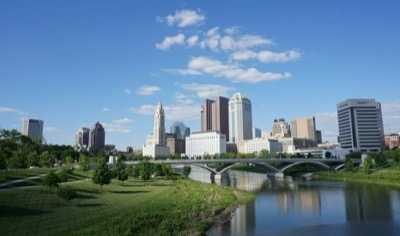 The downtown Columbus, Ohio skyline on a clear day, with LeVeque Tower rising above the Scioto River and a bridge along the Scioto Mile