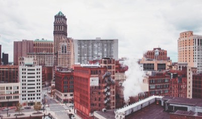 An elevated view of downtown Detroit, with the green copper crown of Book Tower rising above red brick buildings and a plume of rooftop steam