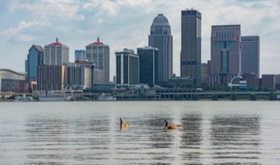 The downtown Louisville, Kentucky skyline seen across the Ohio River on a bright day, with the domed 400 West Market tower, riverboats along the waterfront, and two geese on the water