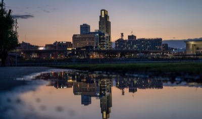 The downtown Omaha, Nebraska skyline at dusk, with the First National Bank Tower lit up and the buildings reflected in a puddle in the foreground